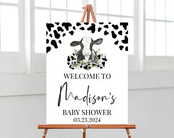 May include: A rectangular white sign featuring a black and white cow print border at the top, a watercolor illustration of a cow surrounded by green leaves, and the text: WELCOME TO Madison's BABY SHOWER 03.23.2024, displayed on a wooden easel.