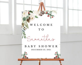 May include: A white sign on a wooden easel with the words "WELCOME TO Samantha's BABY SHOWER DECEMBER 23, 2021." The sign is decorated with watercolor floral arrangements of white flowers, green leaves, and red berries.