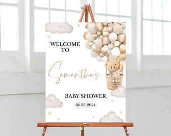 May include: Welcome to Samantha's Baby Shower sign with a teddy bear in a hot air balloon made of gold and white balloons. The sign has a white background with gold stars and clouds.