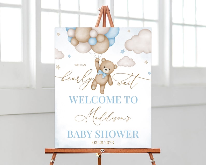 May include: A baby shower sign on an easel features a teddy bear holding balloons. The sign reads "We can bearly wait" and "Welcome to Maddyson's Baby Shower 03.28.2023" in blue and gold lettering, with watercolor clouds and stars.