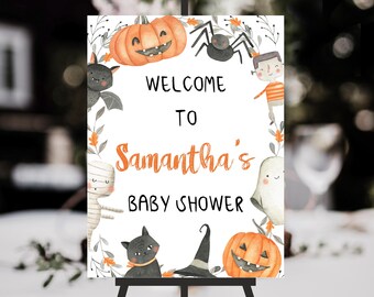 May include: A rectangular sign with a white background featuring the text "WELCOME TO Samantha's BABY SHOWER" in black and orange. The border is decorated with watercolor illustrations of pumpkins, a spider, a bat, a mummy, a ghost, and a cat.
