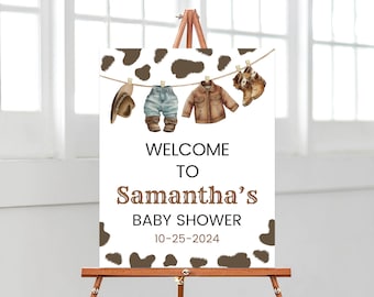 May include: Welcome to Samantha's Baby Shower sign with a brown and white cow print background. A string of clothes hangs from clothespins, including a cowboy hat, jeans, a jacket, and boots.