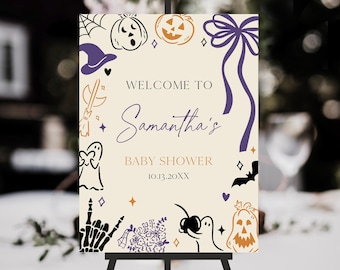 May include: A Halloween-themed baby shower welcome sign on an easel. The sign features a cream background with purple and orange illustrations of pumpkins, bats, and ghosts. Text reads "Welcome to Samantha's Baby Shower 10.13.20XX."