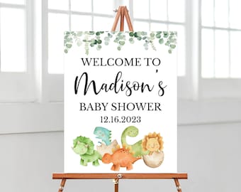 Könnte beinhalten: Ein rechteckiges, weißes Willkommensschild für eine Babyparty mit grünen Eukalyptusblättern am oberen Rand, schwarzem Text "WELCOME TO Madison's BABY SHOWER 12.16.2023" und einer bunten Aquarellillustration von fünf kleinen Dinosauriern unten.