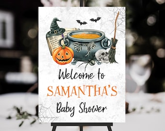 May include: A Halloween-themed baby shower sign featuring a cauldron, pumpkin, skull, and bats. The sign reads "Welcome to SAMANTHA'S Baby Shower" in black and orange lettering. A witch's hat and broom are also depicted.