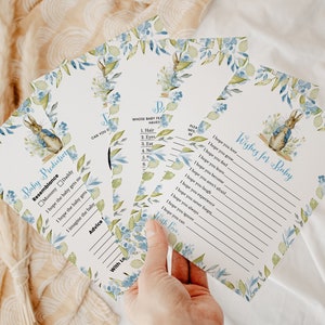 May include: A set of baby shower game cards featuring a watercolor illustration of Peter Rabbit. The cards have blue and green floral accents and include the text "Baby Predictions" and "Wishes for Baby".