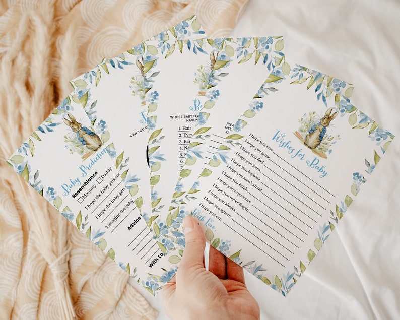 May include: A set of baby shower game cards featuring a watercolor illustration of Peter Rabbit. The cards have blue and green floral accents and include the text "Baby Predictions" and "Wishes for Baby".
