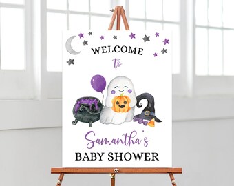 May include: A white sign on an easel with a Halloween baby shower theme. The sign features a ghost holding a pumpkin, a cauldron, a witch's hat, and the text "Welcome to Samantha's Baby Shower" in black and purple.