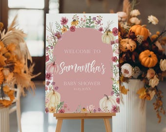 May include: A baby shower welcome sign on an easel. The sign has a pink background with an arch shape. The text reads "Welcome to Samantha's Baby Shower" with the date below. Pumpkins and floral arrangements decorate the sign.