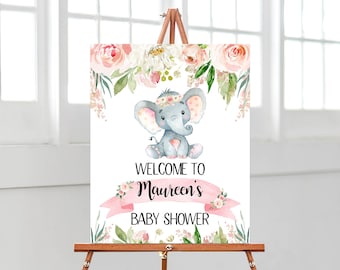 May include: A welcome sign for a baby shower with a pink and green floral design. A gray elephant with a pink flower crown is in the center of the sign. The text on the sign reads "Welcome to Maureen's Baby Shower".