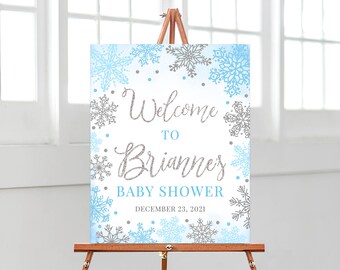 Puede incluir: Un letrero de bienvenida para baby shower sobre un caballete de madera. El letrero presenta un tema invernal con copos de nieve azules y grises y las palabras "Bienvenida a la baby shower de Brianne" en una fuente brillante. También se muestra la fecha "23 de diciembre de 2021".