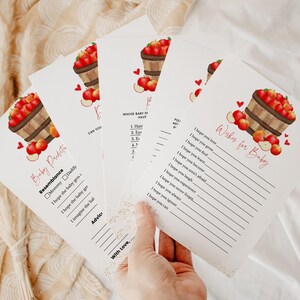 May include: Baby shower game cards with apple basket illustrations. The cards feature text such as "Baby Predictions" and "Wishes for Baby". The cards are white with red and brown accents.