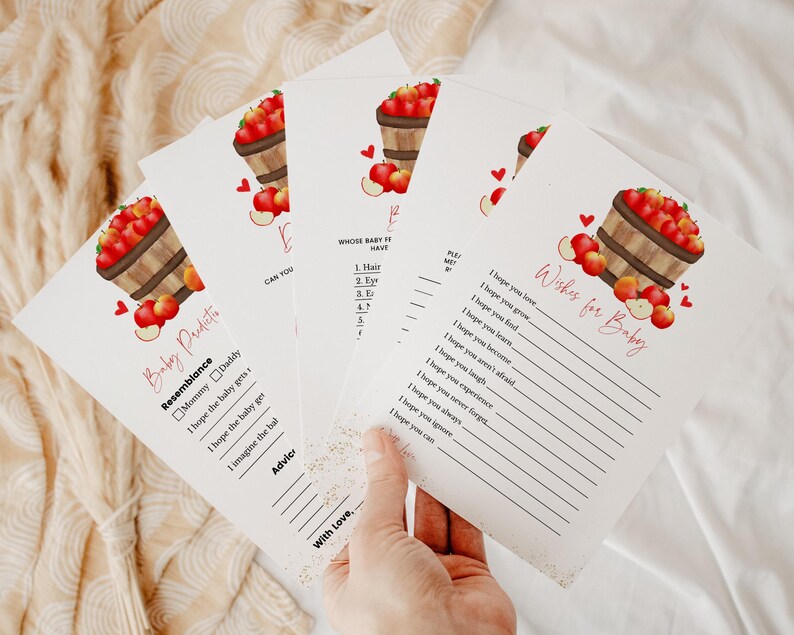 May include: Baby shower game cards with apple basket illustrations. The cards feature text such as "Baby Predictions" and "Wishes for Baby". The cards are white with red and brown accents.