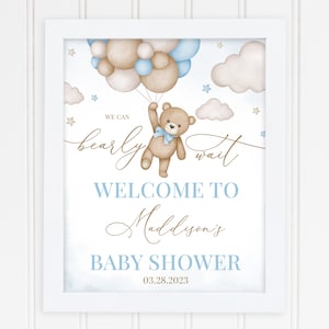 May include: A framed baby shower sign with a watercolor design. The sign features a teddy bear holding balloons, with the text "We can bearly wait" and "Welcome to Maddyson's Baby Shower" in blue and gold script. The date is 03.28.2023.