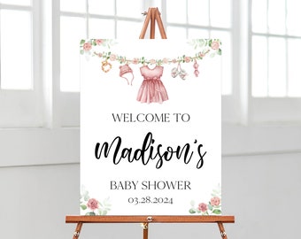 May include: A baby shower welcome sign displayed on an easel. The sign reads "Welcome to Madison's Baby Shower 03.28.2024". The top of the sign features a watercolour illustration of baby clothes and floral decorations.