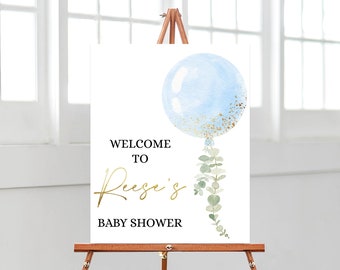 Puede incluir: Un letrero blanco sobre un caballete de madera con el texto "WELCOME TO Reese's BABY SHOWER" en letras negras y doradas. Un globo de acuarela azul claro con detalles dorados y vegetación cuelga de la parte inferior del globo.