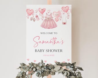 May include: A pink and white baby shower sign with the text "Welcome to Samantha's Baby Shower 02-14-2025". The sign is decorated with pink hearts, a pink dress, and other baby shower decorations.