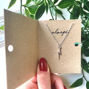 Puede incluir: Un collar de plata con un dije de rayo y la palabra "always" escrita en una tarjeta marrón.