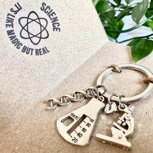 Può includere: Un portachiavi in metallo color argento con un ciondolo a forma di elica di DNA, un ciondolo a forma di becher da 100 ml, un piccolo ciondolo circolare con la lettera S e un ciondolo a forma di microscopio. Il testo stampato recita: Science, it's like magic but real.