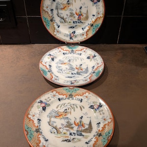 Op de afbeelding: Drie antieke decoratieve borden met een Aziatisch geïnspireerd ontwerp. Elk bord heeft een centrale scène met figuren, bomen en gebouwen, omgeven door een bloemen en geometrische rand in oranje, blauw en groen.