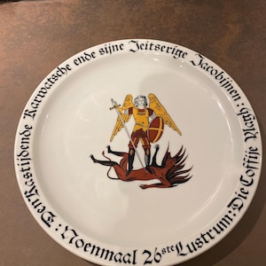 Puede incluir: Plato de cerámica blanca con una ilustración central de una figura con armadura dorada con espada y escudo, de pie sobre un demonio caído. Texto negro rodea el borde. El plato tiene un fondo marrón.