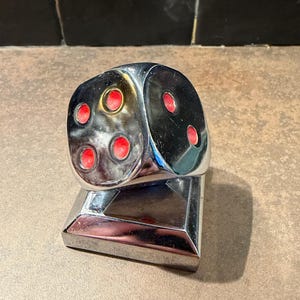 May include: A chrome dice-shaped ornament with red dots, mounted on a rectangular base. The dice shows the numbers 1, 2, and 3. The object has a reflective surface, possibly a decorative item or paperweight.