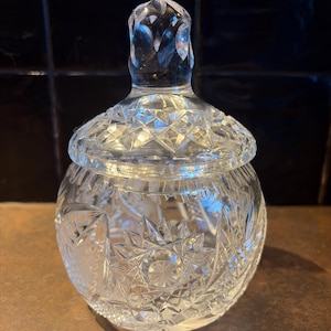 Op de afbeelding: Een heldere, geslepen glazen pot met een deksel. De pot heeft een ronde body en een deksel met een knop. Het glas is ingewikkeld gesneden met geometrische patronen, waardoor een sprankelend effect ontstaat. De pot wordt waarschijnlijk gebruikt voor het bewaren van kleine items of als decoratief stuk.