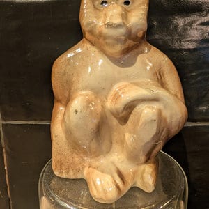 Op de afbeelding: Een keramieken apenbeeldje in zittende positie, met een lichtbruine glazuur en donkerbruine accenten. De aap heeft een gedetailleerd gezicht en rust op een heldere glazen pot. De achtergrond is een donker, getextureerd oppervlak.