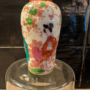 Può includere: Un piccolo vaso decorativo in ceramica con base bianca, caratterizzato da un design di geisha e motivi floreali in rosso, verde e rosa. Il bordo è arancione. Il vaso è esposto su una superficie di vetro trasparente.