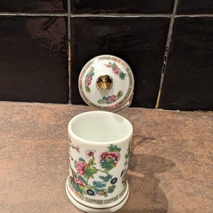 Op de afbeelding: Een witte keramische container met bijpassende deksel. De container en deksel zijn versierd met een bloemenpatroon in roze, groen en blauw. De deksel heeft een goudkleurige knop. De container is cilindrisch en heeft een licht uitlopende basis.