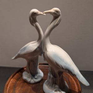 Puede incluir: Dos figuras de pájaros de porcelana blanca y azul claro, enfrentadas. Los pájaros tienen cuellos y picos largos. Están montados sobre una base de madera tallada de color marrón oscuro.