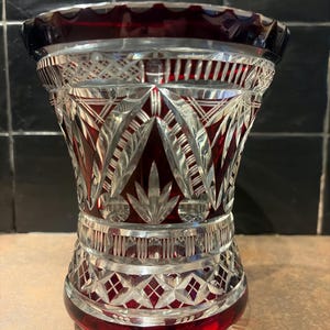 Op de afbeelding: Een decoratieve vaas van geslepen glas in robijnrood en helder. De vaas heeft gedetailleerde geometrische en bladachtige patronen. De rand en de basis zijn dieprood, met een heldere body, wat een contrast creëert. Een decoratief stuk.