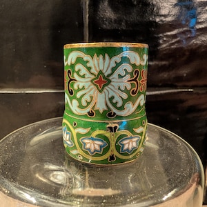 Op de afbeelding: Een kleine, cilindrische decoratieve container met een scharnierend deksel. De container heeft een groene basis en bovenkant met bloempatronen in wit, blauw, rood en zwart. De rand is afgewerkt met goud. De container staat op een glazen oppervlak.