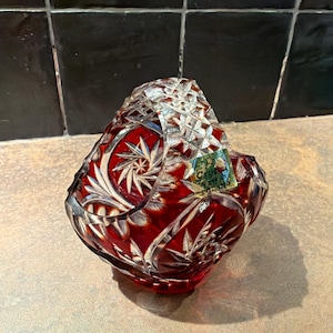 Op de afbeelding: Een robijnrode kristallen mand met een heldere, geslepen glazen handgreep. De mand heeft ingewikkelde patronen van sterren en bladeren. Aan de zijkant is een kleine sticker zichtbaar. De mand wordt waarschijnlijk gebruikt voor decoratieve doeleinden.