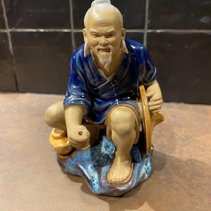 Op de afbeelding: Een keramieken beeldje van een oudere Aziatische man in een blauwe jurk, zittend met een hoed en een kleine container. Het beeldje heeft een witte baard en haar en staat op een blauw-bruine basis.