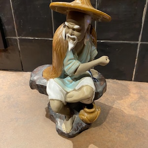 Op de afbeelding: Een keramieken beeldje van een oude man met een strohoed, zittend op een rots. De figuur heeft een lange witte baard en lang haar. Hij is gekleed in lichtblauwe en witte kleding, met een kleine bruine pot naast zich.