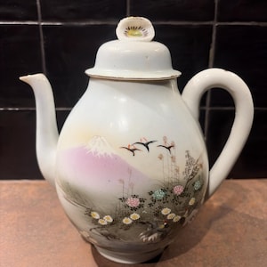 Op de afbeelding: Witte keramische theepot met een geschilderde Japanse landschapsscène. Het ontwerp toont een roze berg, vliegende vogels en bloemmotieven. Het deksel is bekroond met een kleine, decoratieve bloem. De theepot heeft een gebogen handvat en een tuit.