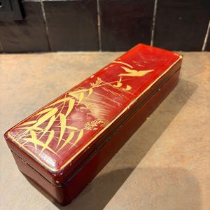 Può includere: Una scatola rettangolare laccata rossa con un disegno dorato di bambù e uccelli. La scatola è lunga e stretta, con un coperchio incernierato. Il design dorato è dettagliato e intricato, aggiungendo un tocco di eleganza alla scatola.