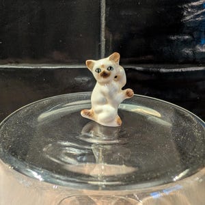 Op de afbeelding: Een kleine, witte keramische kattenfiguur met bruine oren en poten. De kat staat in een speelse pose, met één poot omhoog. Het beeldje heeft groene ogen en een bruine neus. De kat zit op een heldere glazen ondergrond.