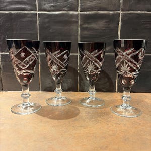 Op de afbeelding: Vier robijnkleurige kristallen wijnglazen met ingewikkelde gesneden patronen. Elk glas heeft een heldere steel en voet, met een gedetailleerd ontwerp van sterren en geometrische vormen. De glazen zijn in een rij gerangschikt.