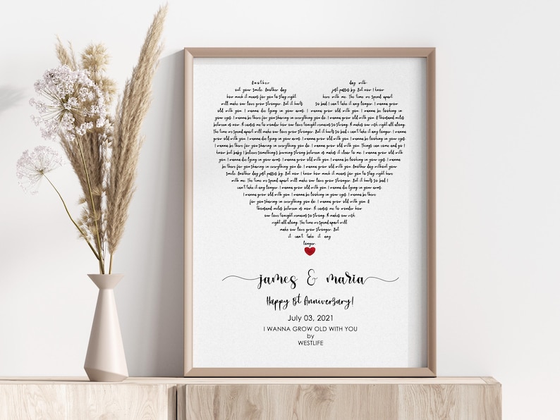 May include: A framed print with a heart-shaped design filled with song lyrics. The print includes the names "James & Maria", the text "Happy 1st Anniversary!", and the date "July 03, 2021". A red heart is at the bottom.