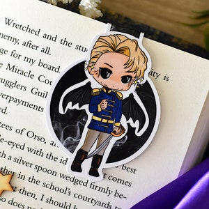 Puede incluir: Una pegatina con un personaje de dibujos animados con cabello rubio, que lleva un uniforme azul y dorado, alas negras y sostiene una espada. El personaje está de pie sobre un fondo negro con humo blanco.