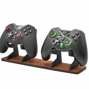 May include: Two black video game controllers are displayed on a wooden stand. The controller on the left has red buttons, while the controller on the right has green buttons.