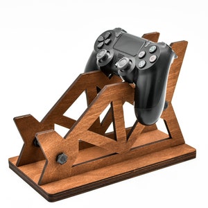 May include: A wooden stand with a geometric design holds a black video game controller. The stand is made of dark brown wood and has a triangular pattern.