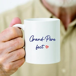 Peut inclure: Mug en céramique blanche avec les mots "Grand-Père fect" en écriture bleue et un cœur rouge. La tasse a une anse standard et est tenue par une personne.