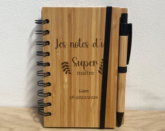 May include: A wooden notebook with a black elastic band and a black pen. The notebook has the text "Les notes du Super maître Liam CP-2023/2024" engraved on the front.