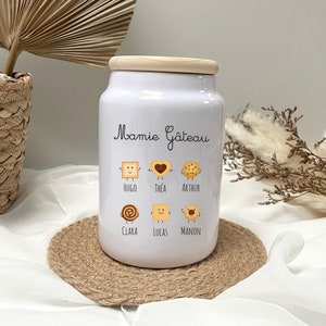 Peut inclure: Une jarre à biscuits en céramique blanche avec un couvercle en bois. La jarre porte l'inscription "Mamie Gâteau" et présente des biscuits de dessin animé avec des noms comme Hugo, Thea, Arthur, Clara, Lucas et Manon.