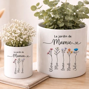 Peut inclure: Deux pots de fleurs en céramique blanche avec des motifs floraux et du texte. L'un dit "Le jardin de Maman" et l'autre "Le jardin de Mamie". Les pots contiennent des plantes et sont posés sur une surface en bois.