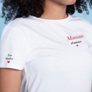 Peut inclure: T-shirt blanc avec le texte "Maman d'amour" en rouge sur le devant et "Léo Ambre" en noir sur la manche gauche. Les deux phrases sont accompagnées d'un petit cœur rouge.