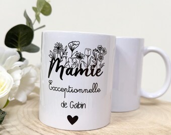 May include: White ceramic mug with a floral design and the word "Mamie" in black script. Below the word "Mamie" is the text "Exceptionnelle de Gabin" and a black heart. A second white mug is behind the first.